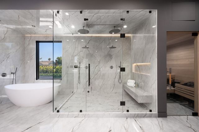 A freestanding tub and large glass-enclosed shower sit beside a timber-lined sauna amid pale veined stone.