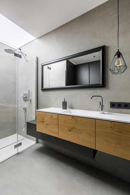 A timber vanity and wide mirror line a grey bathroom beside a walk-in shower and pendant lighting.