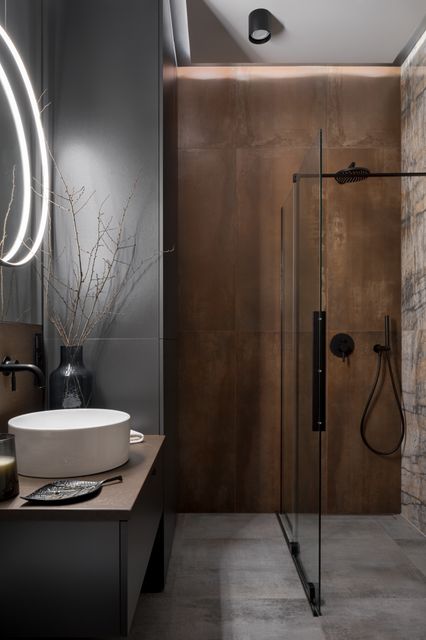 A vessel basin and glass shower stand beside dark grey surfaces and a warm, rust-toned feature wall.