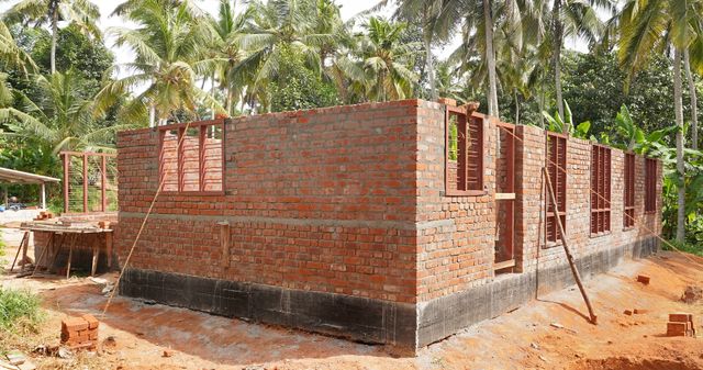Unfinished red-brick walls and metal window frames form a house shell surrounded by palms and construction materials.