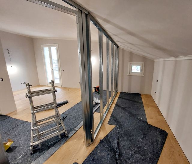 Exposed metal studs form an interior partition beneath a sloping ceiling, with a ladder and protective floor coverings nearby.