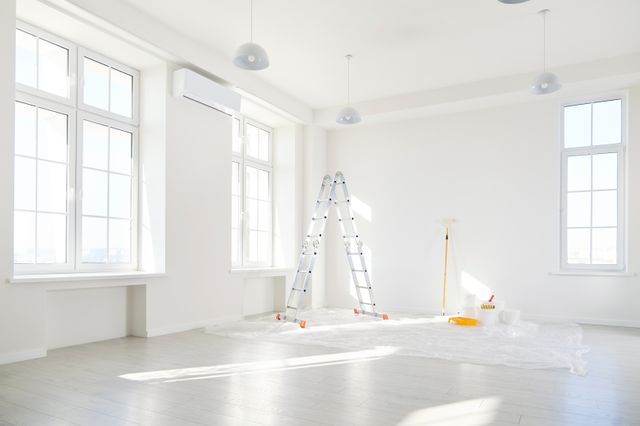 A ladder, paint rollers and buckets stand on protective sheeting in a bright room with white walls and tall windows.