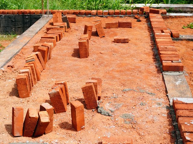 Red bricks are arranged along a raised plinth, with single brick courses marking wall lines and reinforcement bars rising at a corner.