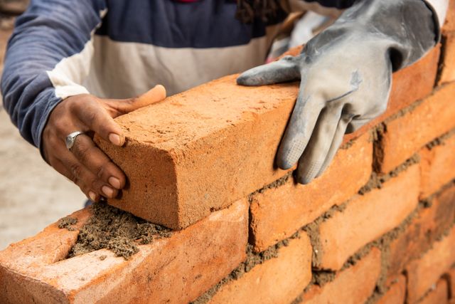A close view of a worker's hands positioning a red brick on a fresh mortar bed above a partially built wall.