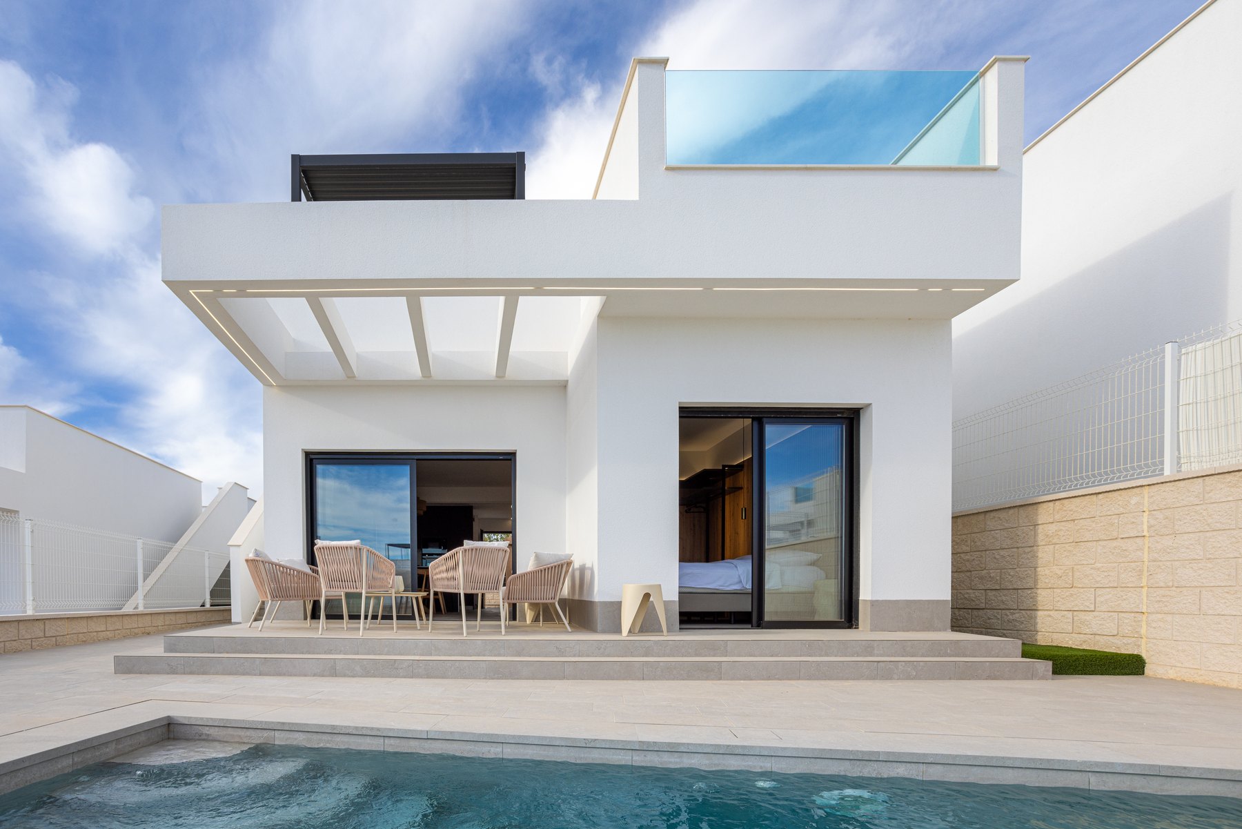 A white villa with dark sliding doors, outdoor seating and an upper glass balustrade overlooks a pool and paved terrace.
