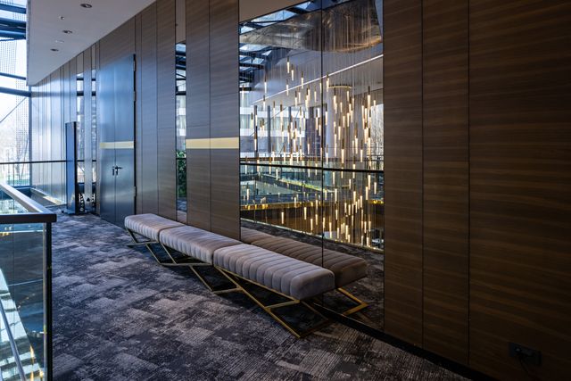 Upholstered benches line dark timber panels and mirrors beside a glass balustrade overlooking suspended pendant lights.