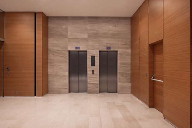 Two metal lift doors sit within a textured stone wall, framed by timber paneling and a pale stone floor.