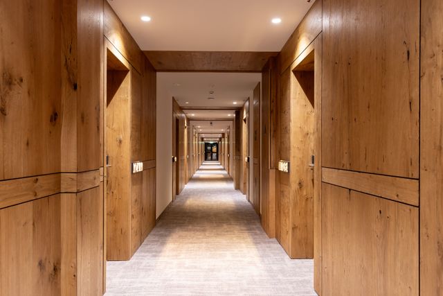 A long carpeted hotel corridor is framed by timber wall panels and repeated guest-room doors under recessed ceiling lights.
