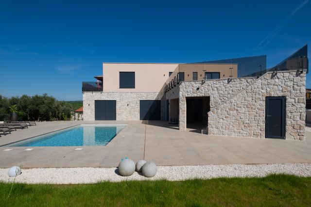 A flat-roofed villa with stone-clad walls, dark shutters and glass balustrades stands beside a rectangular pool and paved terrace.