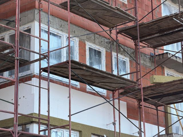 Metal scaffolding and timber platforms stand in front of brick walls, covered windows and partly installed exterior insulation panels.