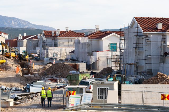 Rows of tiled-roof houses under construction stand behind scaffolding, earthworks and excavators, with two workers walking through the site.