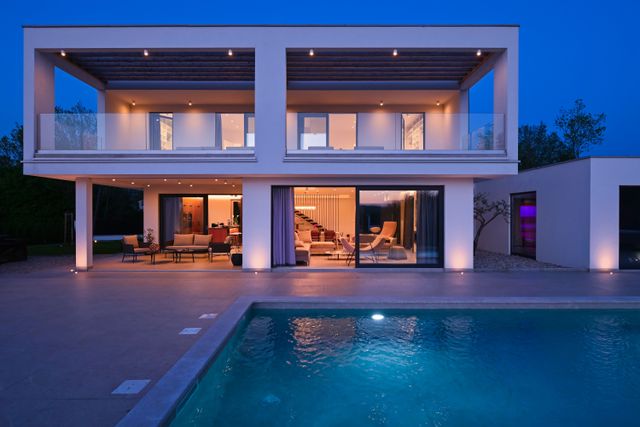 Warm interior lights glow through a white two-storey villa with covered balconies, glass balustrades and a swimming pool in front.