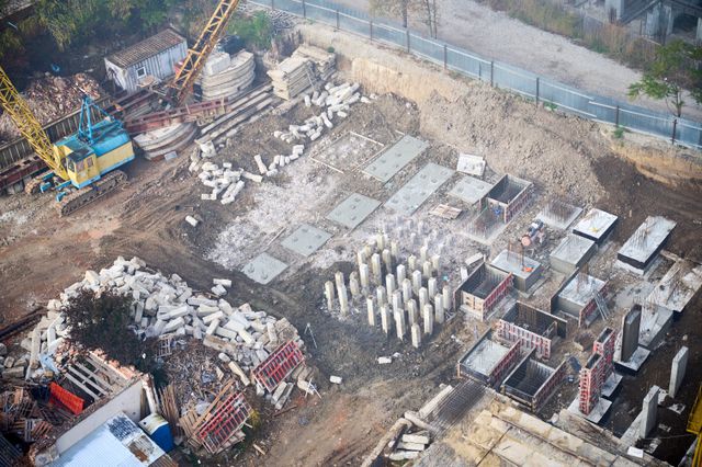 An elevated view shows concrete piles, reinforced bases and formwork across an excavated site beside a tracked crane and stacked materials.