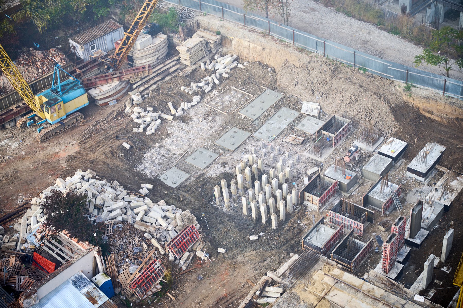 An elevated view shows concrete piles, reinforced bases and formwork across an excavated site beside a tracked crane and stacked materials.