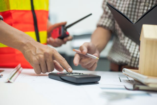 Cropped hands use a calculator over drawings while another person holds a pen, with a radio, folder and small house model nearby.
