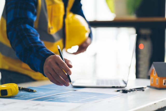 A cropped person points a pencil toward blue drawings on a desk beside a laptop, measuring tape, drawing compass and small house model.