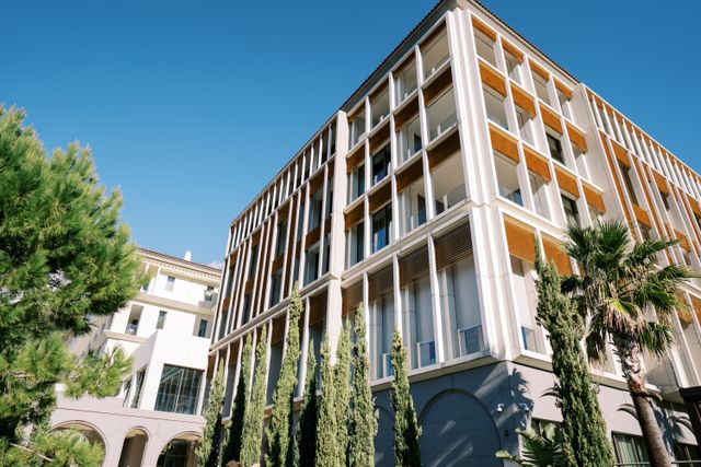 A pale multi-storey hotel has repeated vertical facade elements, glass balcony rails and warm-toned screens, with tall planting in front.