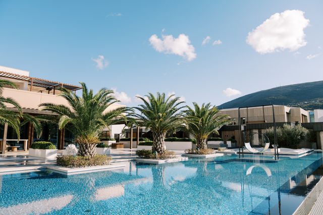 Palm trees and low planted islands border a blue resort pool, with shaded terraces, a suspended seat and a hillside beyond.