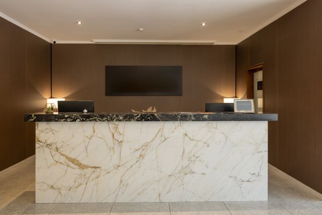 A broad pale veined-stone reception desk with a dark top stands against brown walls with warm lamps on either side.