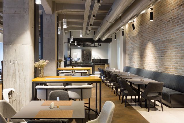 Dining tables and upholstered seating sit between concrete columns and a brick wall, beneath exposed ceiling ducts and pendant lights.