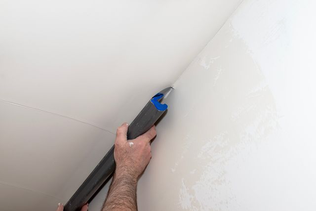 A close view of hands drawing a broad plastering blade across a pale wall near the ceiling junction.