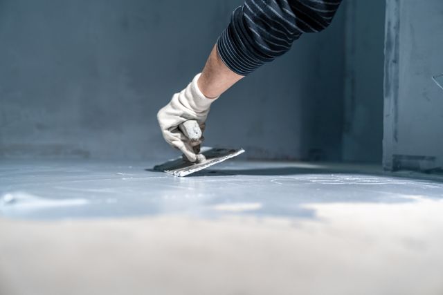 A gloved hand uses a trowel to spread a blue-grey coating across a floor beside a coated wall.
