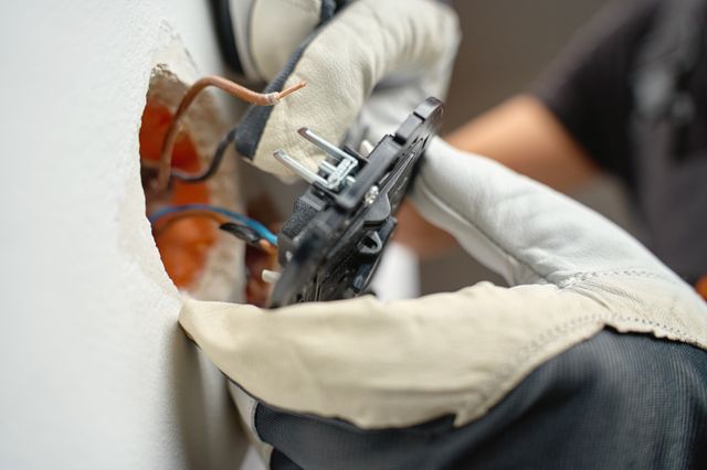 Gloved hands hold an electrical mechanism beside exposed wires and an orange recessed wall box.