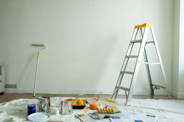 A stepladder and long-handled roller stand beside a pale wall, with paint tins, trays and brushes on protective floor sheets.