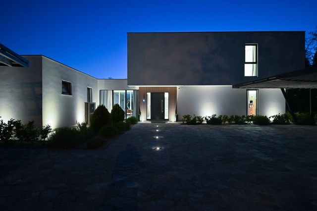 Ground lights lead across a paved forecourt toward a modern villa entrance, with planted borders, dark upper walls and lit windows.