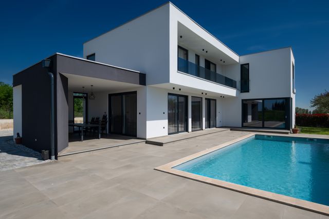 A white two-storey villa with dark-framed glazing, an upper glass balcony and a shaded dining terrace overlooks a rectangular pool.