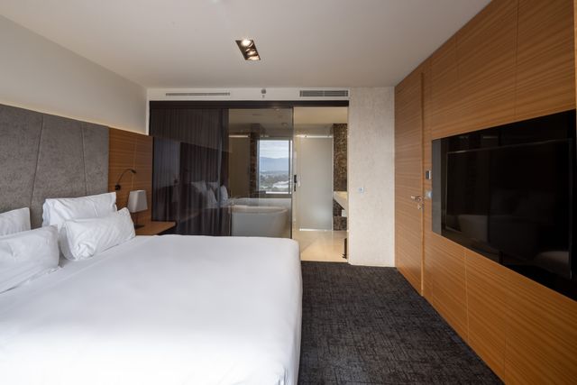 A hotel bedroom combines white bedding, an upholstered headboard and timber wall panels, with a glass partition revealing the bathroom beyond.