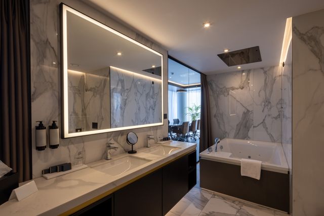 Double basins, a wide illuminated mirror and a bathtub sit within a hotel bathroom lined with pale veined wall and floor surfaces.