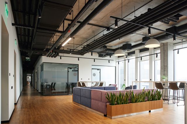 Soft lounge seating and raised window counters sit beside glass meeting rooms under an exposed ceiling with dark pipework.