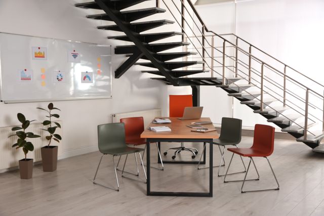 A wood-topped meeting table with red and green chairs sits beneath an open staircase beside a wall-mounted whiteboard.