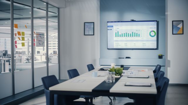 A long pale meeting table and dark chairs face a wall display in a glass-partitioned room overlooking workstations.