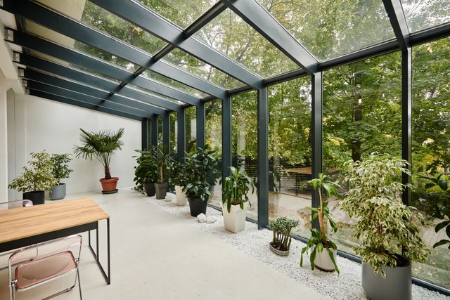 A glazed roof and tall windows surround potted plants and a simple table with translucent chairs.