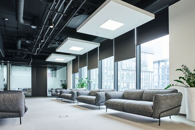 Rows of grey sofas sit beneath suspended white light panels beside tall shaded windows and potted plants.