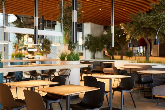 Pale cafe tables and dark chairs sit beneath slim pendant lights, with planted dividers and a timber-slat ceiling.