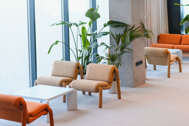 Beige lounge chairs and white coffee tables sit beside leafy plants, with orange seating beyond a broad column.