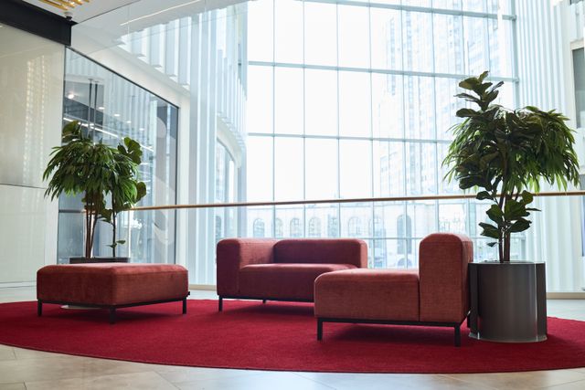 Deep red lounge chairs and a matching ottoman sit on a red rug between tall plants beside full-height windows.