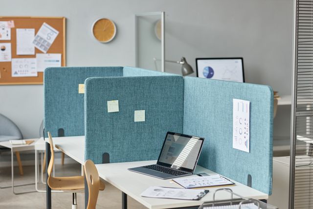 White desks with blue divider panels hold a laptop and printed charts, with curved chairs and a noticeboard nearby.