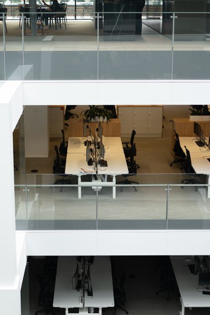 Shared desks and black task chairs sit behind glass railings in a multilevel office, with a seated person on the upper level.