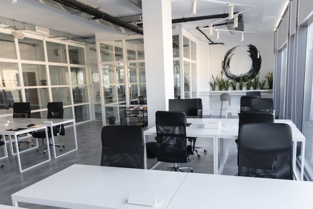 Rows of white desks and black task chairs sit beside tall windows, with glazed rooms and a planted counter at the back.