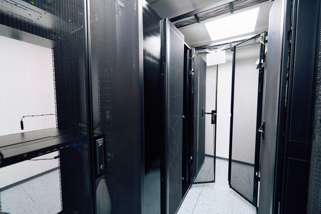 Tall mesh-fronted equipment cabinets line a narrow aisle, with open doors and cables overhead.