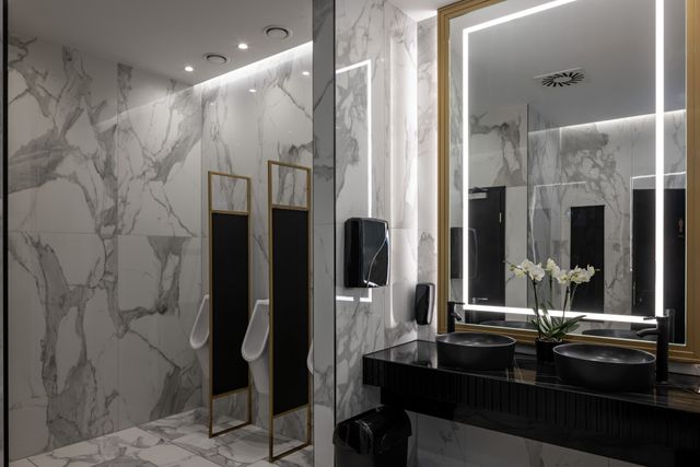 Two black vessel basins sit below an illuminated mirror beside veined wall panels and partitioned urinals.