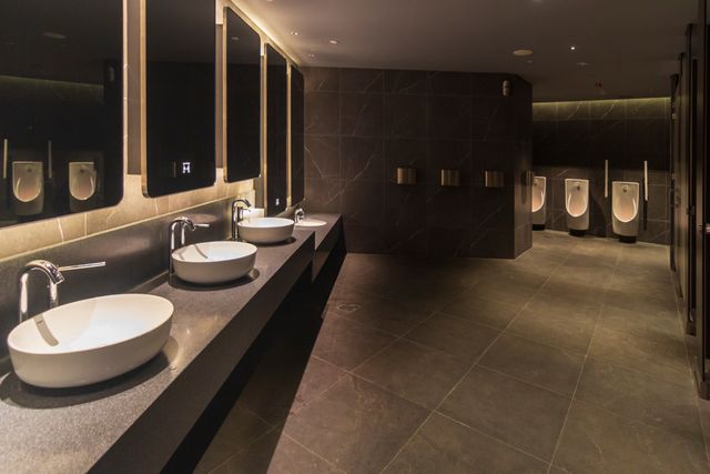 A row of white basins and illuminated mirrors lines a dark-toned restroom, with urinals at the far end.