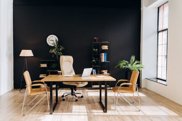 A desk with an open laptop and high-backed chair faces two visitor chairs, with shelving and plants against a dark wall.