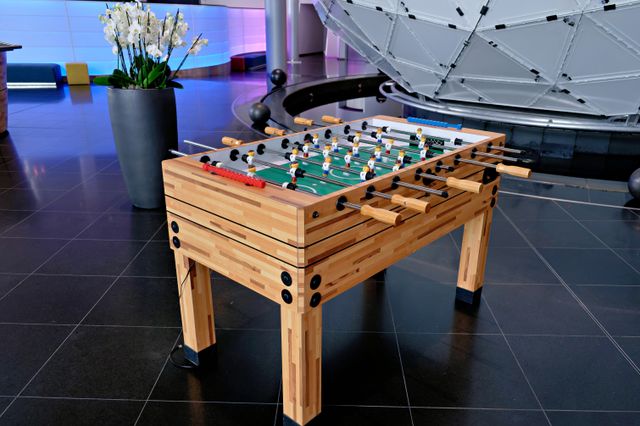 A timber foosball table stands beside a large planter in a brightly lit lobby with dark tiled flooring.
