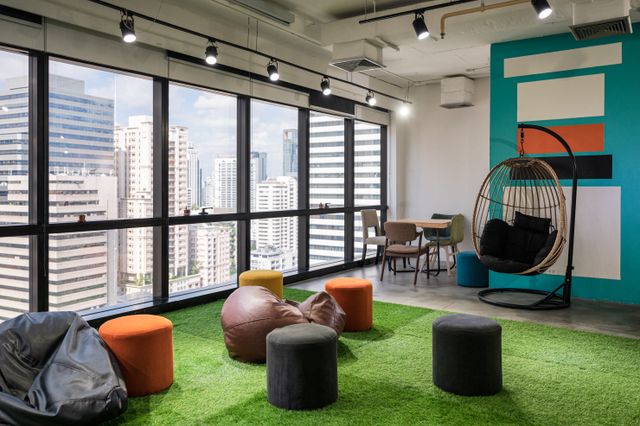 Beanbags and colorful stools share a green floor covering beside a hanging chair and large city-facing windows.