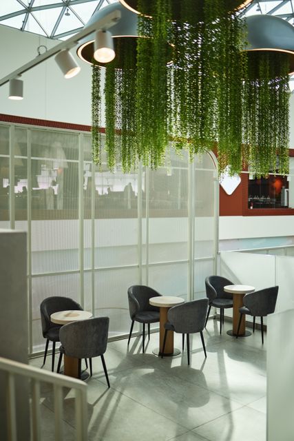 Small round tables and dark upholstered chairs sit beside a pale mesh divider below hanging greenery and ceiling lights.